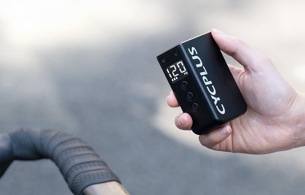 Rowerzysta operujący pompką Cycplus AS2 PRO MAX nad kierownicą szosową z wyraźnym odczytem ciśnienia.